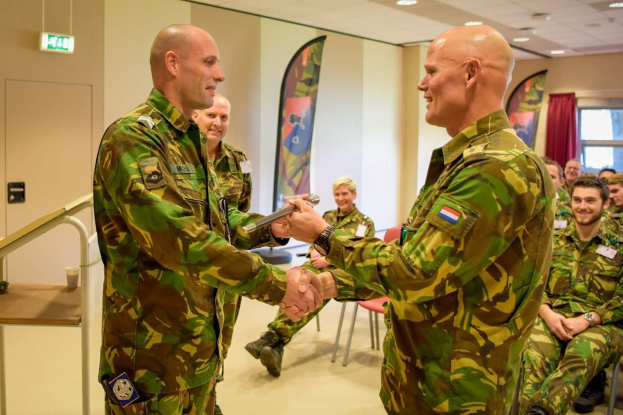 Zwei Männer in deutscher Militäruniform, die sich die Hände schütteln, vor sitzenden Zuschauern an einem formellen Treffen, mit einem Tisch, Bannern und anderen Hintergrundelementen.