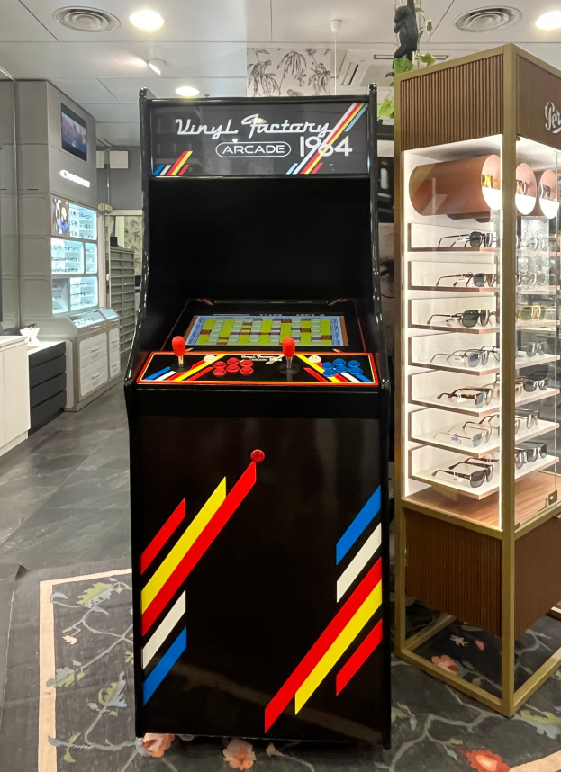 Ein Geschäft mit einem zentralen Retro-Arcade-Automaten, einem rechten Gestell mit Gläsern und Hintergrund-Elementen wie Schränken, einem Fernseher und anderen Gegenständen sowie Deckenleuchten, die eine retro-mäßige Atmosphäre schaffen.