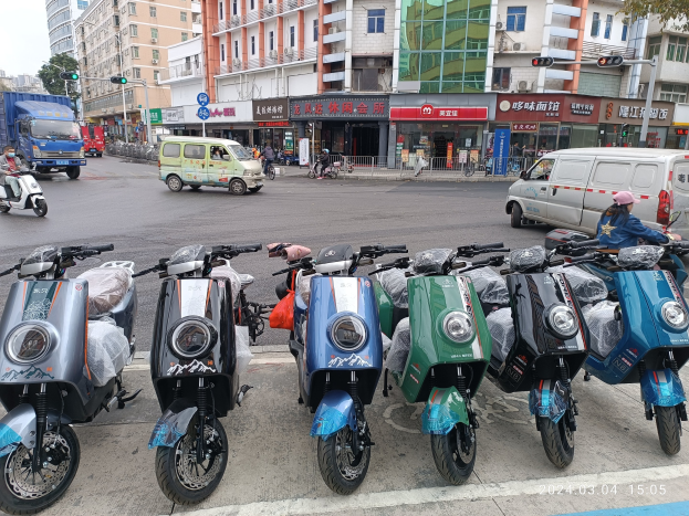 Eine Reihe von Elektro-Scootern, die entlang einer Straße geparkt sind, mit einer Person in einer Mütze davor, Gebäuden, Schildern, Verkehrszeichen, Bäumen und einem klaren Himmel im Hintergrund.
