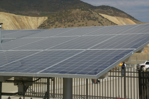 Solarpanels auf dem Dach eines Gebäudes mit Bergen im Hintergrund, ein paar Menschen und Fahrzeuge auf der rechten Seite und ein Geländer unten.
