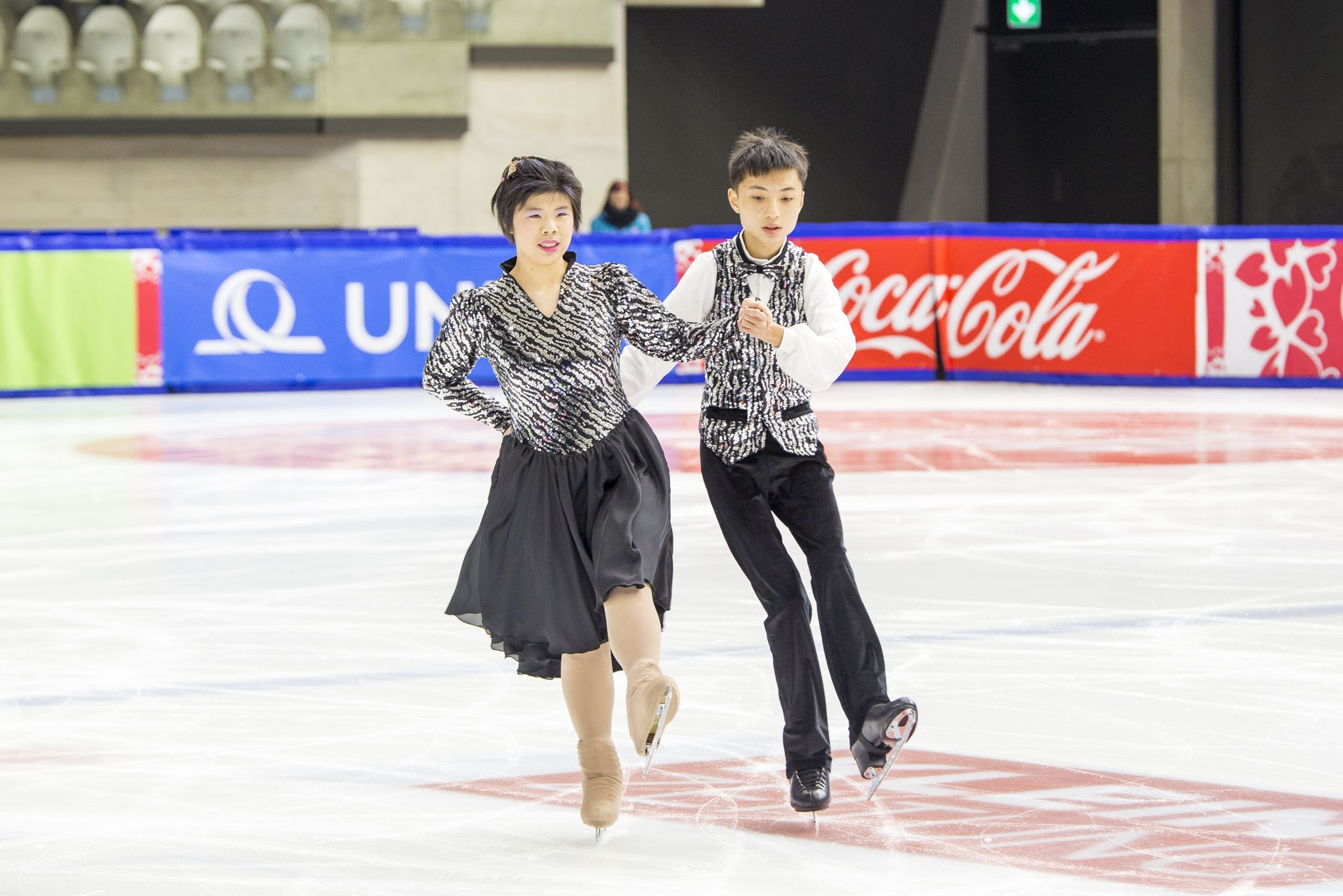 Zwei Eiskunstläufer, ein Mann und eine Frau, gleiten Hand in Hand über eine Eisbahn mit Zuschauern und Absperrungen im Hintergrund.