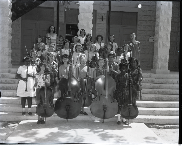 Kinder stehen auf Treppenstufen und halten Musikinstrumente, darunter Celli, Geigen und Trompeten, mit Säulen und einer Wand mit Türen im Hintergrund.