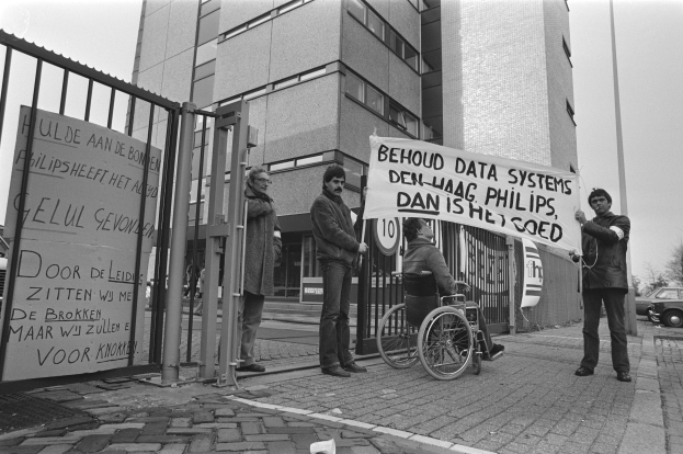 Ein Mann im Rollstuhl hält ein Transparent vor einem Gebäude, umgeben von einer Gruppe von Menschen, einem Metalltor, einem Schild, Fahrzeugen auf der Straße, einem Straßenschild, Bäumen und einem bewölkten Himmel.