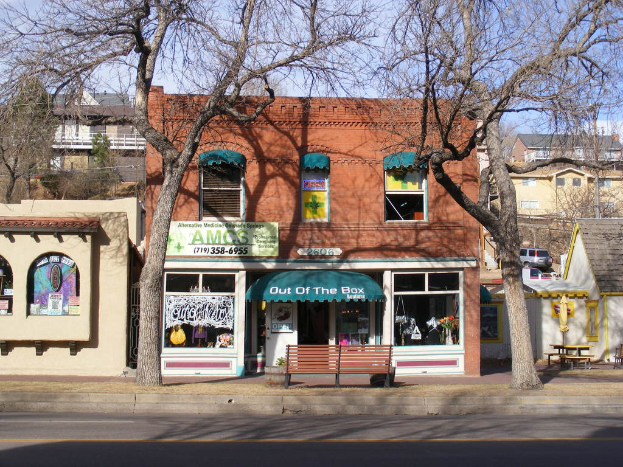 Eine Straße im Vordergrund mit Gebäuden, Bänken, Bäumen, einem Fahrzeug und dem Himmel im Hintergrund.