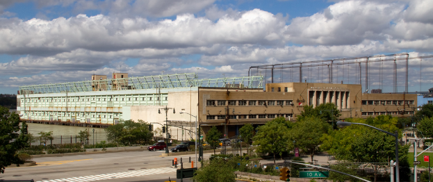 Eine Straße mit Fahrzeugen, Bäumen und Straßenlaternen entlang einer Wasserfläche, die zu einem großen architektonischen Gebäude führt.