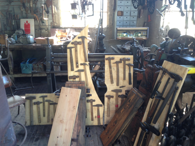 Eine Werkstatt voller verschiedener Werkzeuge, Geräte und Holzplanken, mit einem Stuhl links, Werkzeugen an der Wand und einem Fenster im Hintergrund.