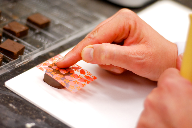Eine Hand, die eine dekorative Abdeckung auf eine Schokolade legt, mit mehr Schokoladen auf einem Tablett links und einem Tisch darunter.
