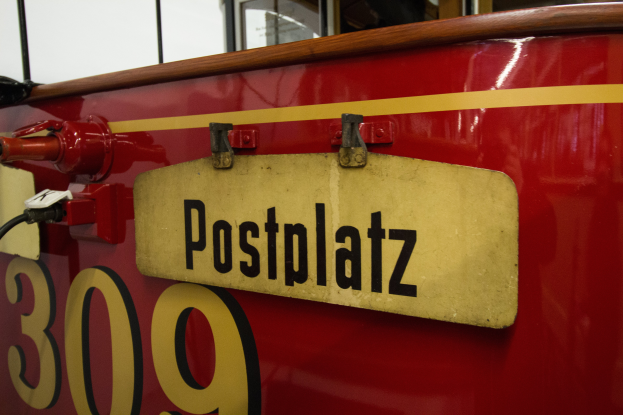 Ein roter und gelber Zug mit 'Postplatz'-Schriftzug und Nummernschild, vor einem Hintergrund aus Glasfenstern und einer Wand.