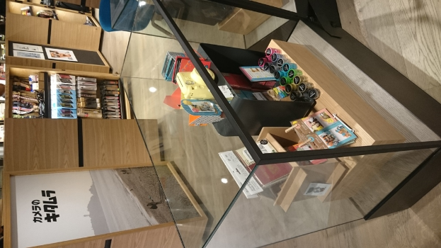 Kartons, Flaschen, Bücher und Poster mit Text sind auf Holzregalen und einem Glastisch angeordnet.