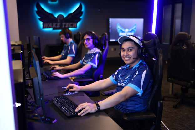 Gruppe von Menschen an Computern mit Headsets, die während eines Esport-Turnieraufbaus mit Monitoren, Tastaturen und einer markierten Hintergrundwand lächeln.