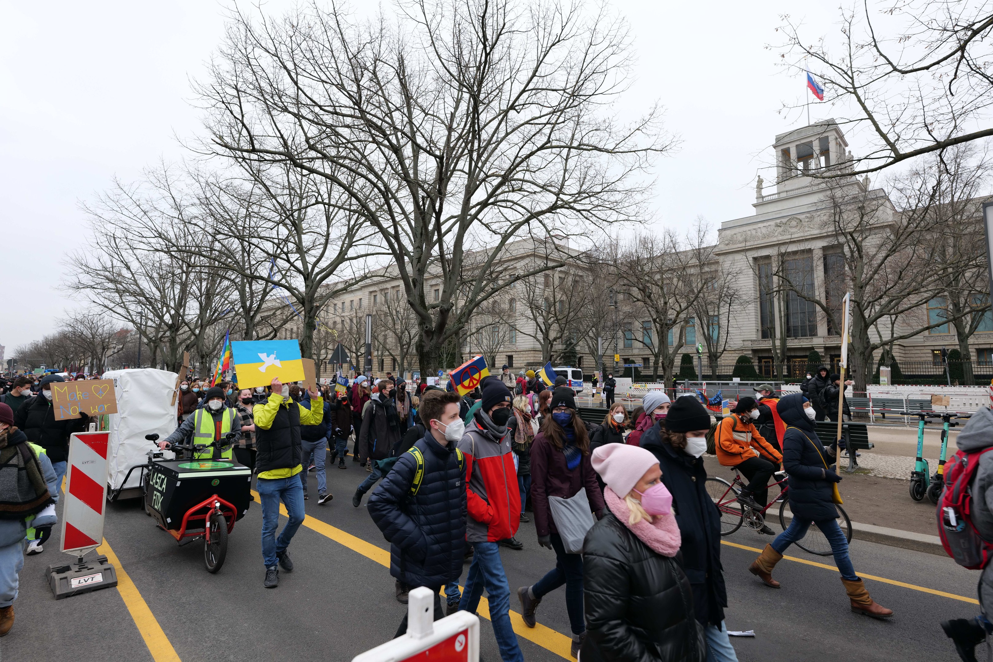Eine große Protestmarsch mit Menschen, die eine Straße in Washington, D.C. entlanggehen, einige halten Schilder und andere fahren Fahrräder, mit Bäumen, Schildern und einem klaren blauen Himmel im Hintergrund.