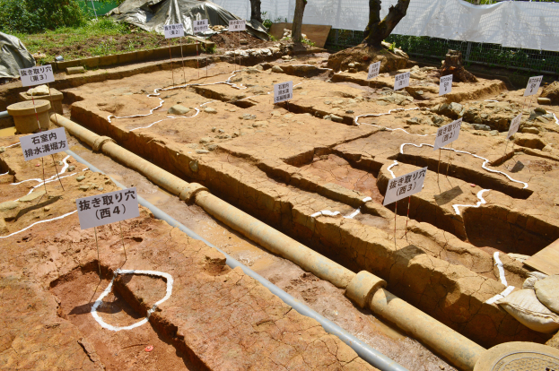 Großes Erdaushubgelände mit Erde, umgeben von Bäumen, Pflanzen, Gras, einem Zaun, einer Abdeckung und einer Wand, mit Tafeln mit Text, Rohren und verstreuten Gegenständen.