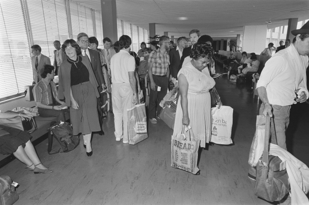 Menschen, die durch einen schwarz-weißen Flughafenterminal gehen und Koffer tragen, Fenster mit Jalousien auf der linken Seite und ein Mann und eine Frau, die in der Mitte stehen und Taschen halten.