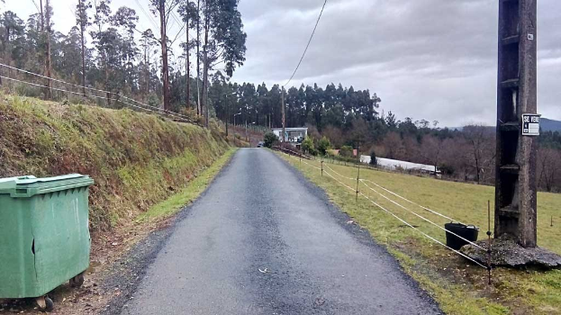 Grüner Müllcontainer am Straßenrand mit Gras, Pflanzen, Bäumen, Pfählen, Drähten und einem Zaun; bewölkter Himmel im Hintergrund.
