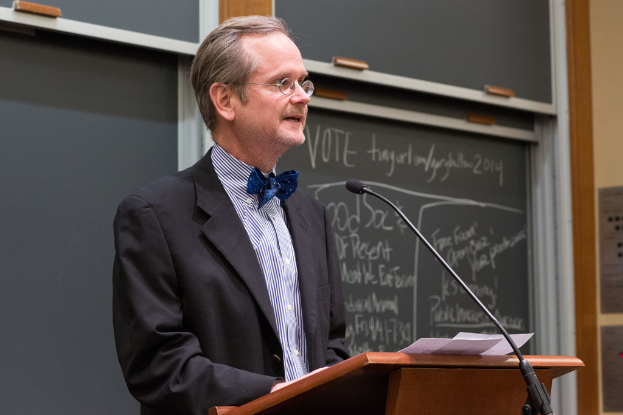 Ein Mann in einem schwarzen Anzug steht an einem Rednerpult und spricht in ein Mikrofon, mit einer Tafel im Hintergrund.
