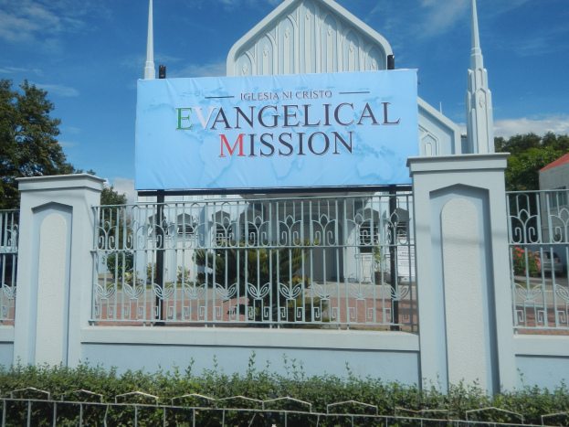 Kirche mit einem Metallzaun, einem "Evangelischen Missions"-Schild, umgeben von Bäumen, einem Haus und Pflanzen unter einem bewölkten Himmel.
