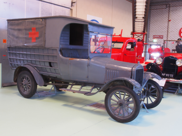 Alter Krankenwagen-Lkw in einem Gebäude mit anderen Fahrzeugen geparkt, ein Schild mit Text links daneben und Hintergrund-Elemente wie Wandbretter, Rohre, Lampen.