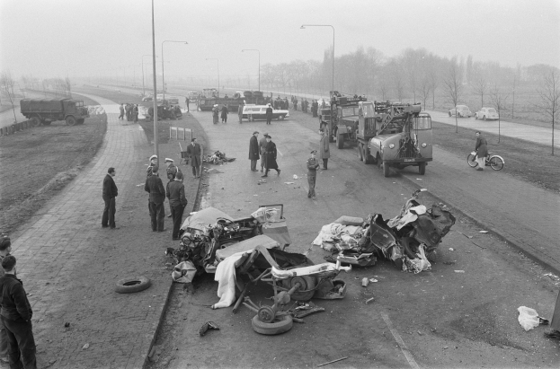 Schwarzes und weißes Bild eines Autounfalls am Straßenrand mit mehreren Fahrzeugen und einer Gruppe von Menschen in der Nähe, Lichtmasten, Bäume und Himmel im Hintergrund.