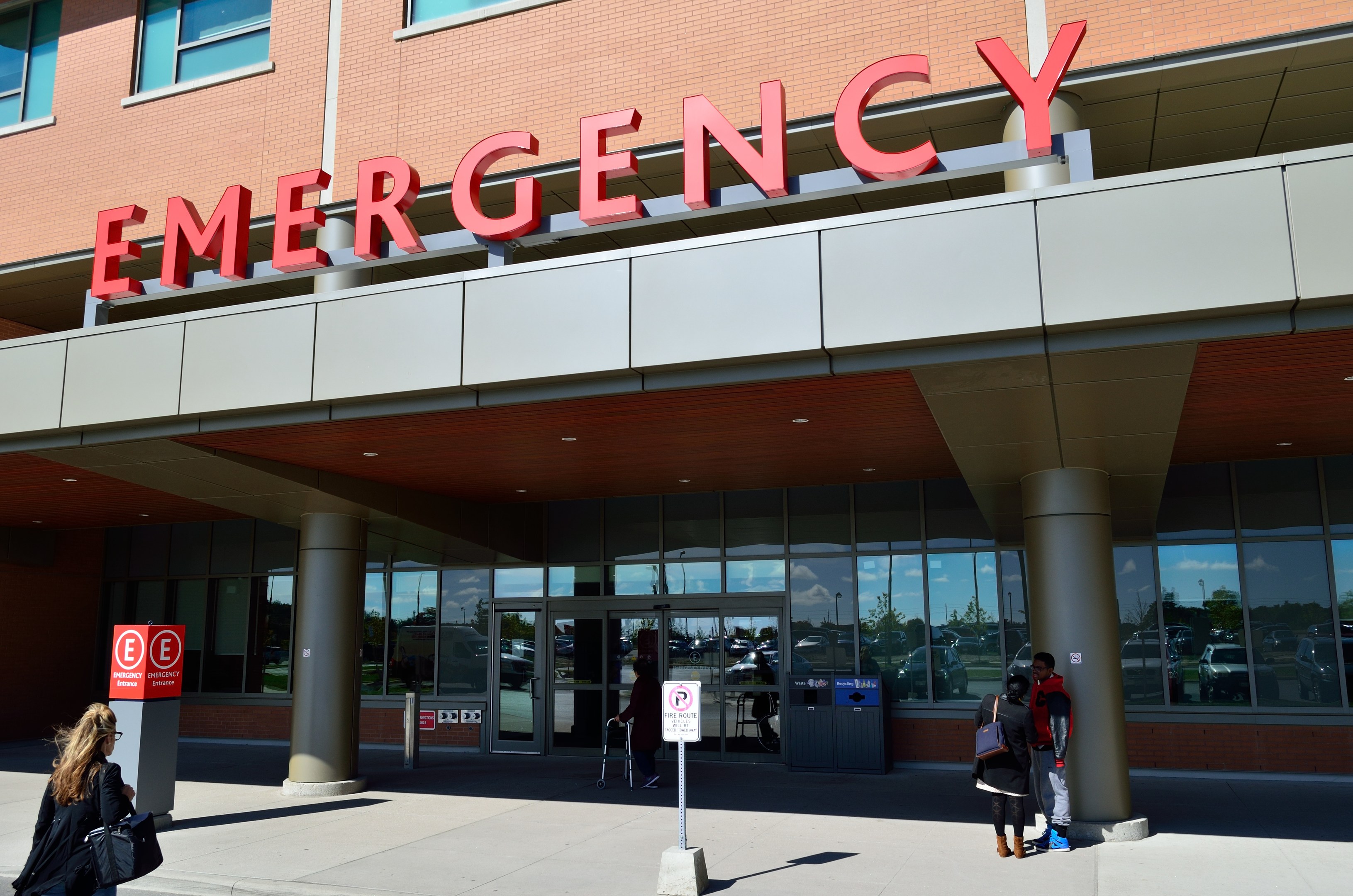 Eine Gruppe von Menschen geht vor einem Glaswand-Notfallgebäude mit Säulen, einem Namensschild, Hinweistafeln auf Pfählen und einem Mülleimer vorbei.