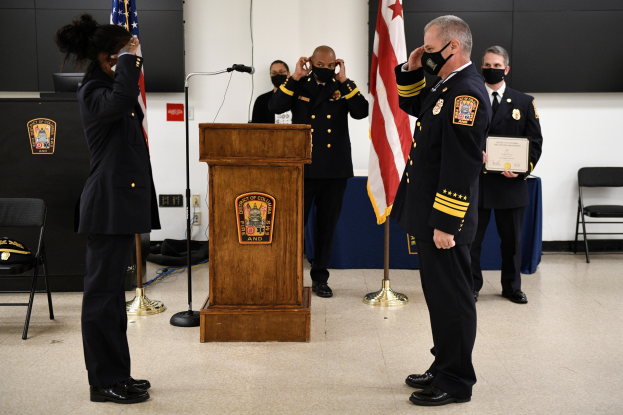 Gruppe uniformierter Personen in Masken vor einem Podium mit Mikrofon, umgeben von Fahnen, Stühlen, Tischen und einer Wand bei einer Vereidigung eines neuen Feuerwehrchefs, wobei eine Person ein Zertifikat hält.