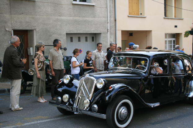 Ein Auto fährt auf einer Straße, auf der links Menschen stehen und im Hintergrund Gebäude zu sehen sind.
