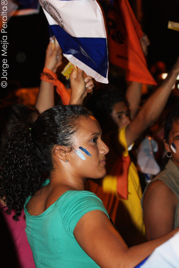 Eine Frau in einem grünen T-Shirt steht bei einer Demonstration und hölt ein weißes Schild, hinter ihr stehen viele andere Menschen.