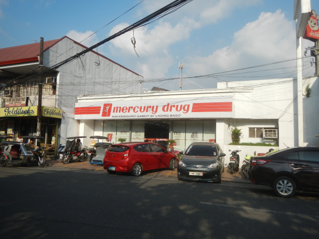 Eine belebte Straßenszene mit Fahrzeugen, Gebäuden mit Schildern, Strommasten mit Drähten und einem bewölkten Himmel, mit einer Mercury Drug Apotheke und parkenden Autos im Vordergrund.