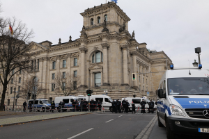 Eine Gruppe von Polizisten vor dem Reichstaggebäude in Berlin, Deutschland, mit Fahrzeugen, einer Absperrung, Verkehrsampeln, Laternenpfählen, Bäumen und Flaggen im Hintergrund.