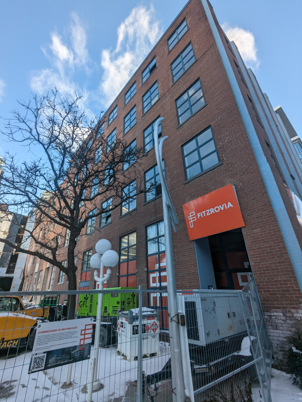 Ein großes Backsteingebäude mit einer Tafel, auf der "Fitzrovia Apartments for Rent in New York City" steht, ist von einem Metallzaun, Straßenlaternen, Bäumen, Fahrzeugen, Pflanzen und einem bewölkten Himmel umgeben.