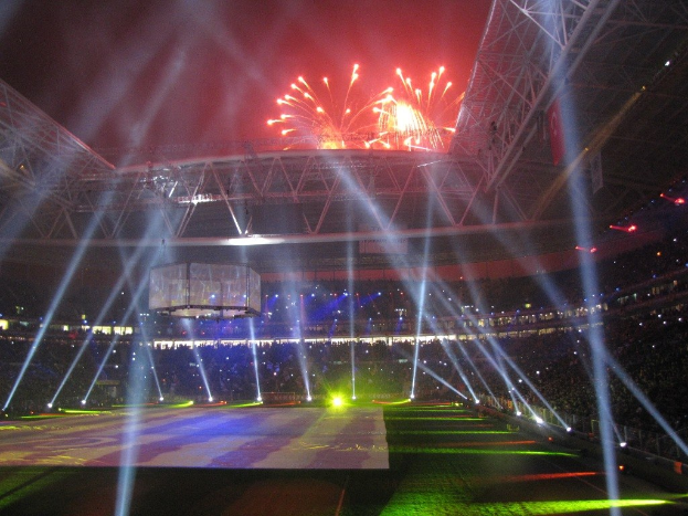 Olympiastadion in London bei Nacht, beleuchtet von hellen Lichtern und Feuerwerken, mit einem großen Bildschirm im Vordergrund und einer Menschenmenge darin.