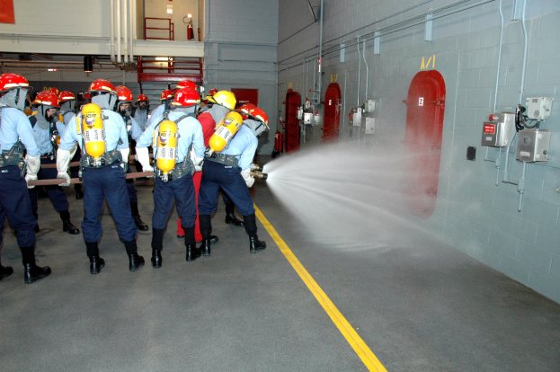 Feuerwehrleute in Helmen und Ausrüstung sprühen sich gegenseitig drinnen in der Nähe einer Wand mit Gegenständen, Rohren, einer Leiter und Feuerlöschern im Hintergrund.