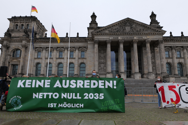 Eine Gruppe von Menschen hält ein Transparent vor dem Reichstagsgebäude in Berlin, Deutschland, das mit Fenstern, Säulen, Bögen und Statuen verziert ist und von Fahnenmasten umgeben ist, mit einem Geländer im Vordergrund und einem Fahrrad auf der rechten Seite, unter einem sichtbaren Himmel.