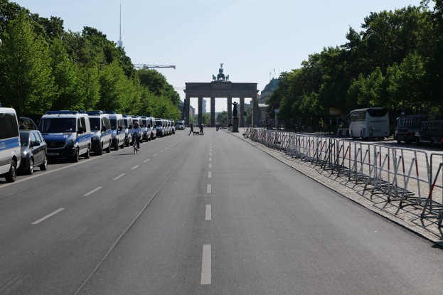 Lange Reihe von Polizeiwagen auf der Seite einer Straße vor dem Brandenburger Tor in Berlin, Deutschland, mit Menschen auf Fahrrädern und auf der Straße stehend, Barrieren, Bäume, ein Bogen mit Statuen im Hintergrund und sichtbarem Himmel.