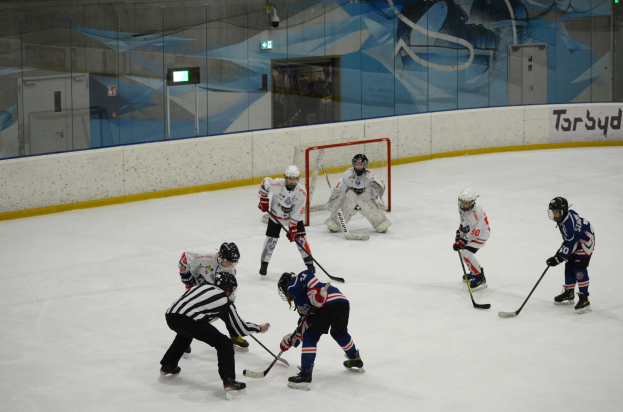 Gruppe von Menschen in Helmen und Hockey-Ausrüstung, die auf einer Indoor-Eisbahn Eishockey spielen, Text an der rechten Wand und ein Gemälde auf der Glaswand im Hintergrund.