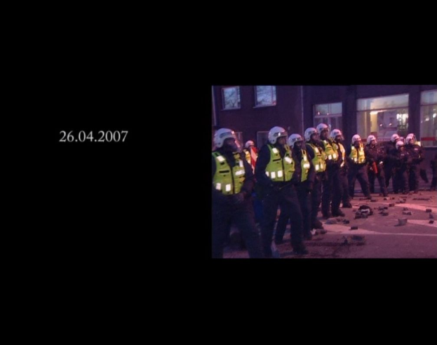 Gruppe von Polizisten in Helmen und Jacken vor einem Gebäude mit Fenstern; Datum auf der linken Seite sichtbar.
