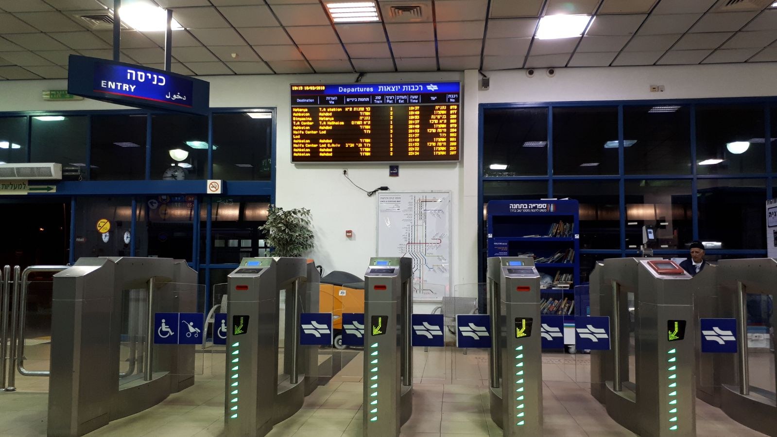 Flughafeneingang mit einem Mann vor einer Metalldrehtür, Metalldetektoren, Informationsschildern, einem Topf mit einer Pflanze, einer Wandtafel, einer Decke mit Leuchten und einem Anzeigebildschirm.