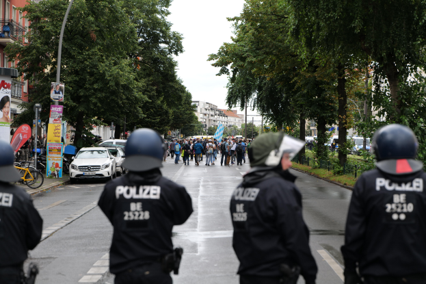 Eine Gruppe von Polizisten in schwarzen Uniformen und Helmen steht an der Straße, mit Menschen, Fahrzeugen, Fahrrädern, Polen, Brettern, Bäumen, Gebäuden und einer Banner mit Text im Hintergrund.