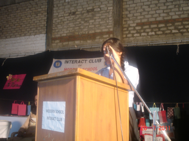 Ein Mädchen in einem weißen Hemd steht neben einem Rednerpult in einem Konferenzraum, mit einem Mikrofonständer rechts daneben, einem weißen Banner an einer Wand hinter ihm und mehreren Taschen an einem Faden rechts.