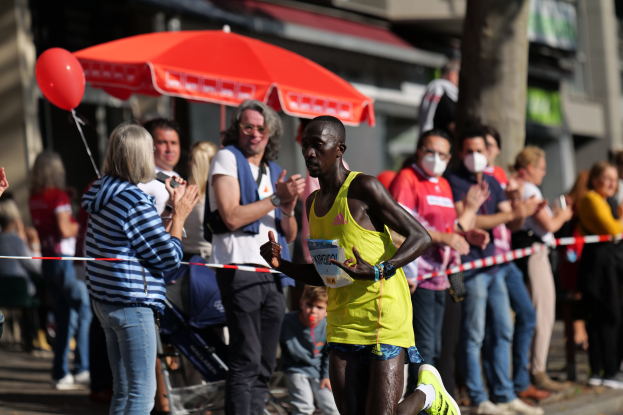 Ein Mann in einem gelben T-Shirt und Laufschuhen sprintet durch einen Marathon, während ihn eine Menge umringt, die klatscht und jubelt, mit einem Schirm und einem Kinderwagen im Hintergrund.