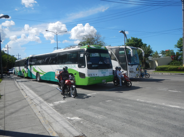 Ein grüner und weißer Shuttlebus steht am Straßenrand mit Motorrädern davor, ein grasbewachsener Fußweg auf der linken Seite und Gebäude, Bäume, Laternenmasten und einen klaren blauen Himmel im Hintergrund.