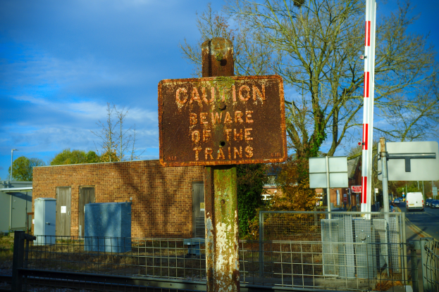 Vorsichtsschild an einem Bahnschienenzaun mit Bäumen, Strommasten, einem Gebäude, Containern, Fahrzeugen auf einer Straße und einem bewölkten Himmel.
