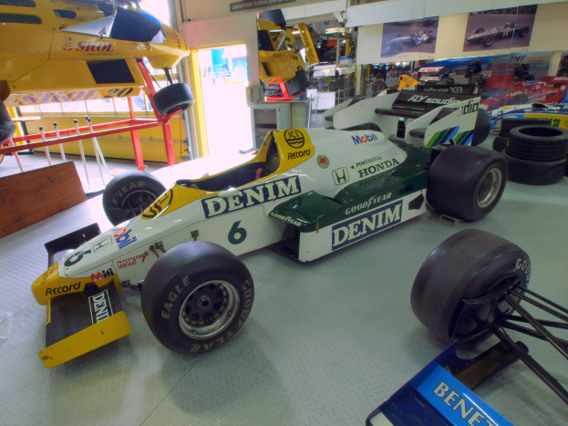 Eine Garage mit verschiedenen Rennwagen, Reifen, einem hölzernen Objekt, einem Geländer, einer Glastür, gerahmten Fotos an der Wand und Deckenleuchten.