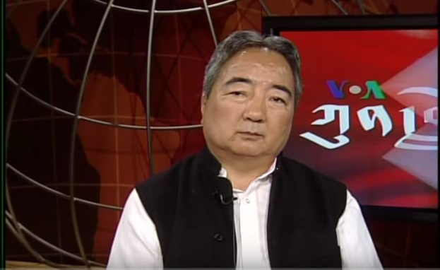 Ein Mann in schwarzer Weste und weißem Hemd sitzt vor einem Fernseher und hält ein Mikrofon, mit einer Weltkugel im Hintergrund, wahrscheinlich beim Diskutieren der Wahlen von 2019.