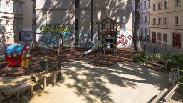 Spielplatz mit Graffiti in einer städtischen Umgebung, umgeben von Gebäuden, Bäumen, Bänken und einer Straße mit Fahrzeugen und Fußgängern unter einem sichtbaren Himmel.