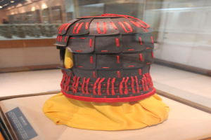 Helm in einer Glasvitrine mit einem gelben Tuch darüber, begleitet von einer Texttafel auf der linken Seite und Deckenlampen im Hintergrund.