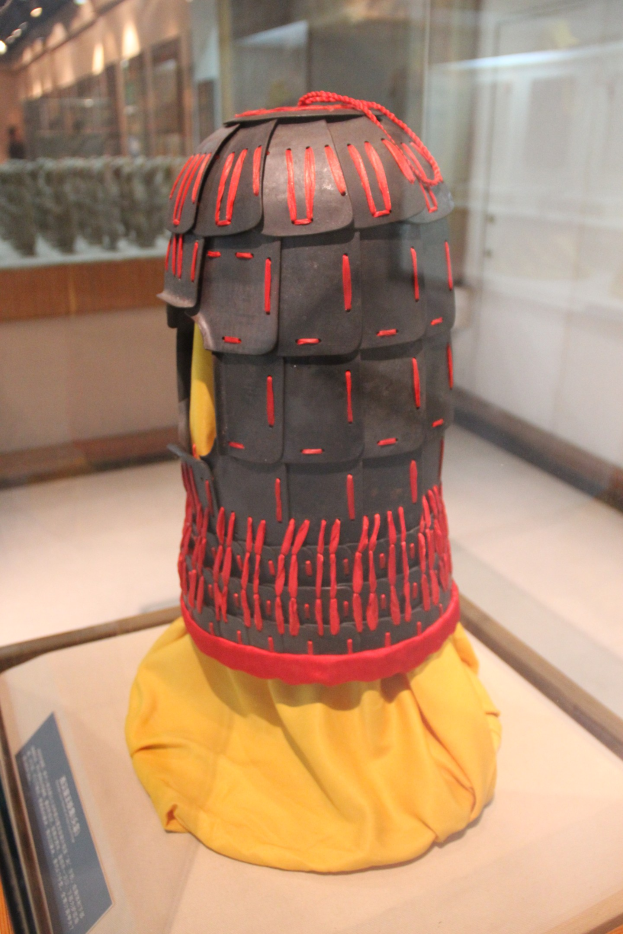 Helm in einer Glasvitrine mit einem gelben Tuch darüber, begleitet von einer Texttafel auf der linken Seite und Deckenlampen im Hintergrund.