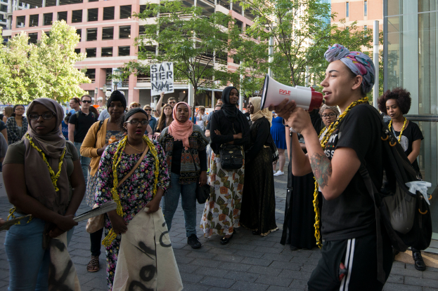 Eine Frau in einem Schal, T-Shirt, Hose und einer Tasche hält einen Lautsprecher und steht zwischen vielen Frauen in Burkas, mit Bäumen, Pfählen, Gebäuden und einer Gruppe von Menschen in der Nähe eines Plakats im Hintergrund.