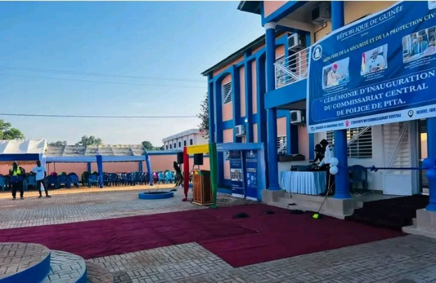 Gruppe von Menschen versammelt vor einem Gebäude mit einem roten Teppich für die Amtseinführung einer neuen Polizeiwache in Guinea, umgeben von Stühlen, Tischen, Bannern, Klimaanlagen, mit Bäumen, Gebäuden und einem klaren blauen Himmel im Hintergrund.