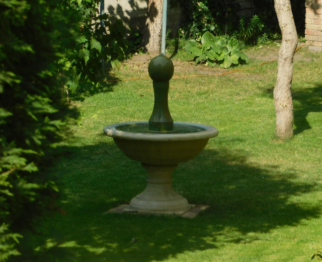 Ein Garten mit einem zentralen Brunnen, umgeben von grünem Gras und Bäumen, und einer Wand mit Pflanzen im Hintergrund.