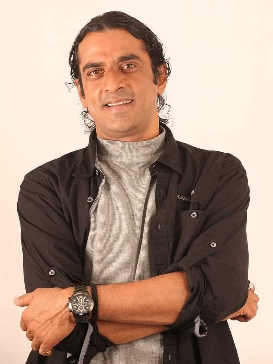 Ein Mann in einem schwarzen und aschfarbenen Kleid mit verschr├Ąnkten Armen, der eine Uhr tr├Ągt und gegen einen wei├čen Hintergrund steht, der nachdenklich erscheint.